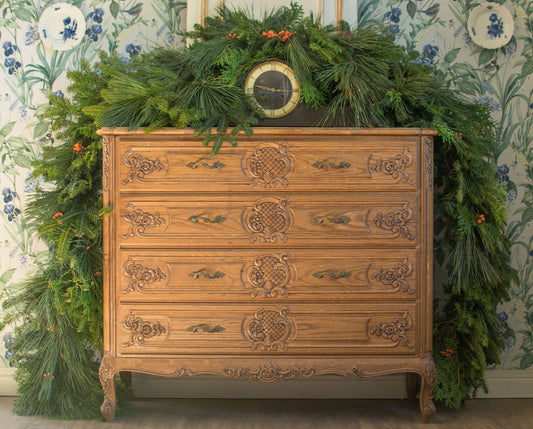 Wooden dresser decorated with a handmade fresh Christmas garland of pine, cedar, and fir branches, set against a floral wallpaper background.