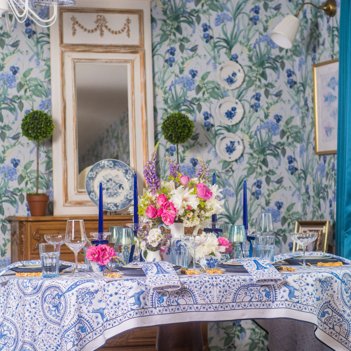Decorative table setting with floral arrangements against a floral wallpapered wall.