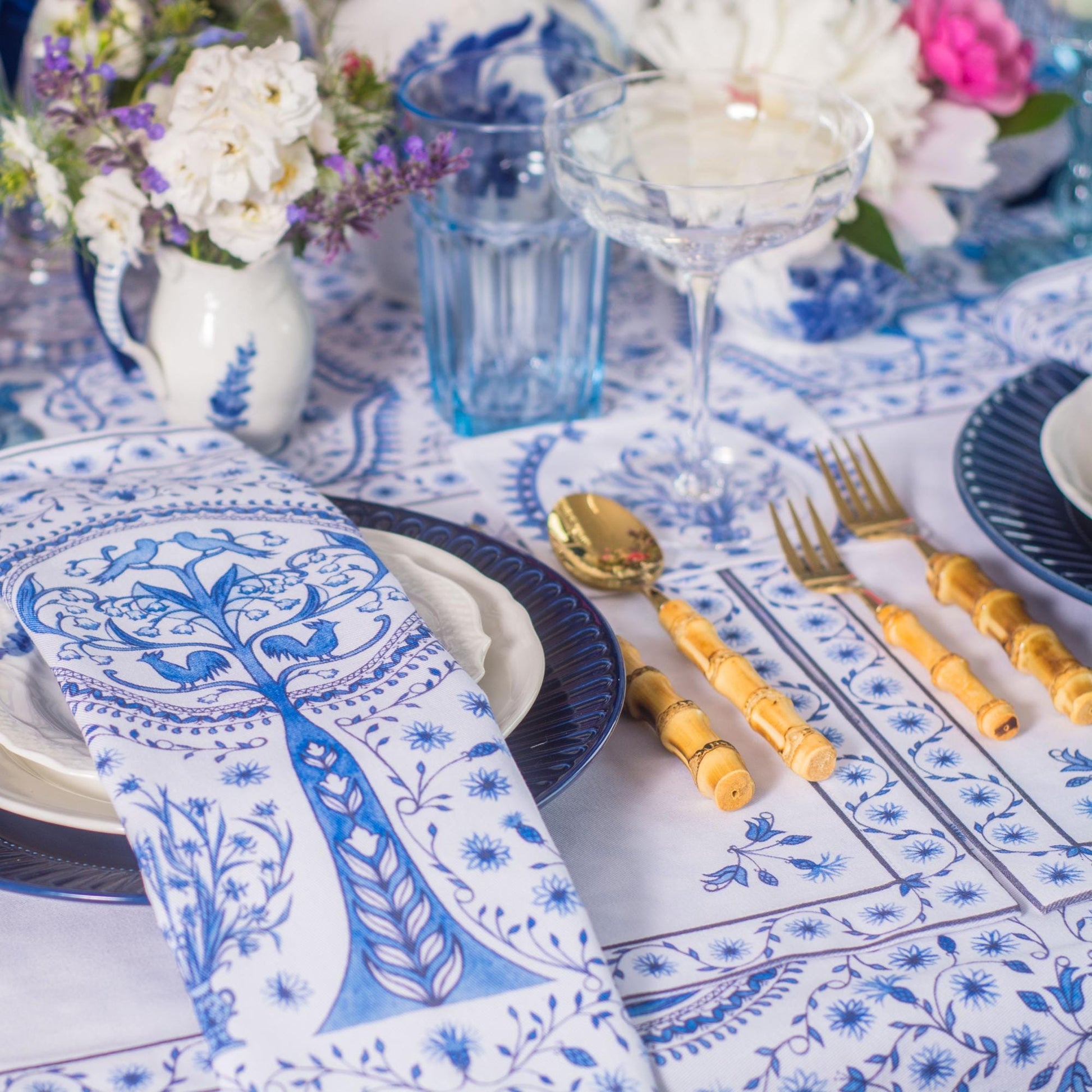 Elegant table setting with blue and white napkin, placemats, floral decorations, and cutlery on a patterned tablecloth.