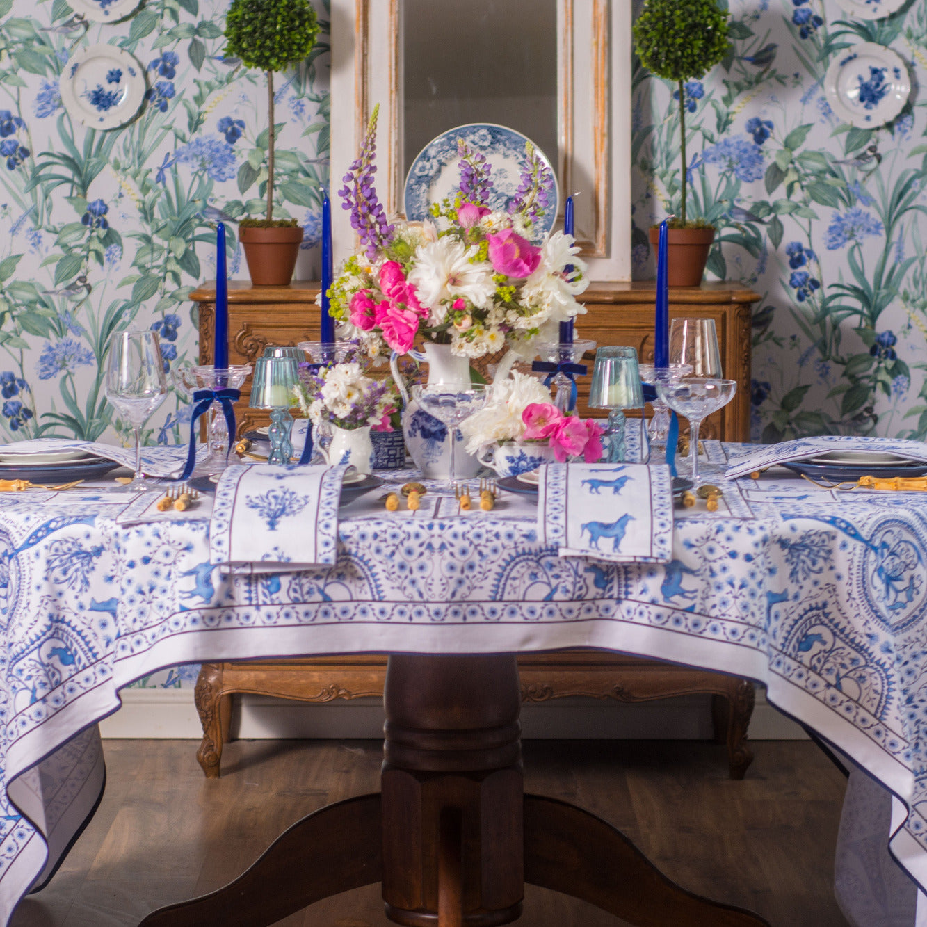 Decorative table setting with floral arrangements and a patterned tablecloth in a room with floral wallpaper.