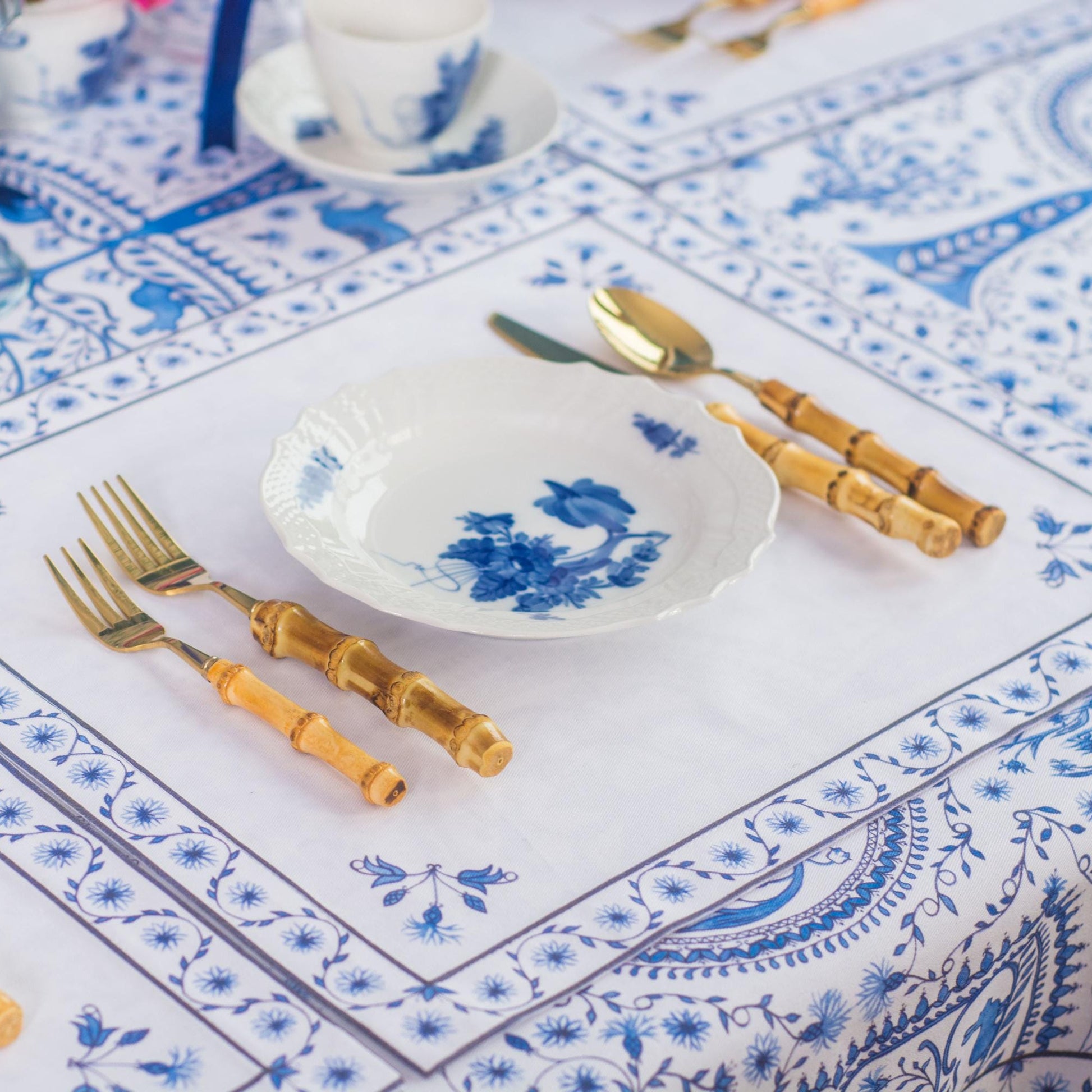 Table setting with blue and white patterned tablecloth, placemats, white plates, gold cutlery, and decorative elements.