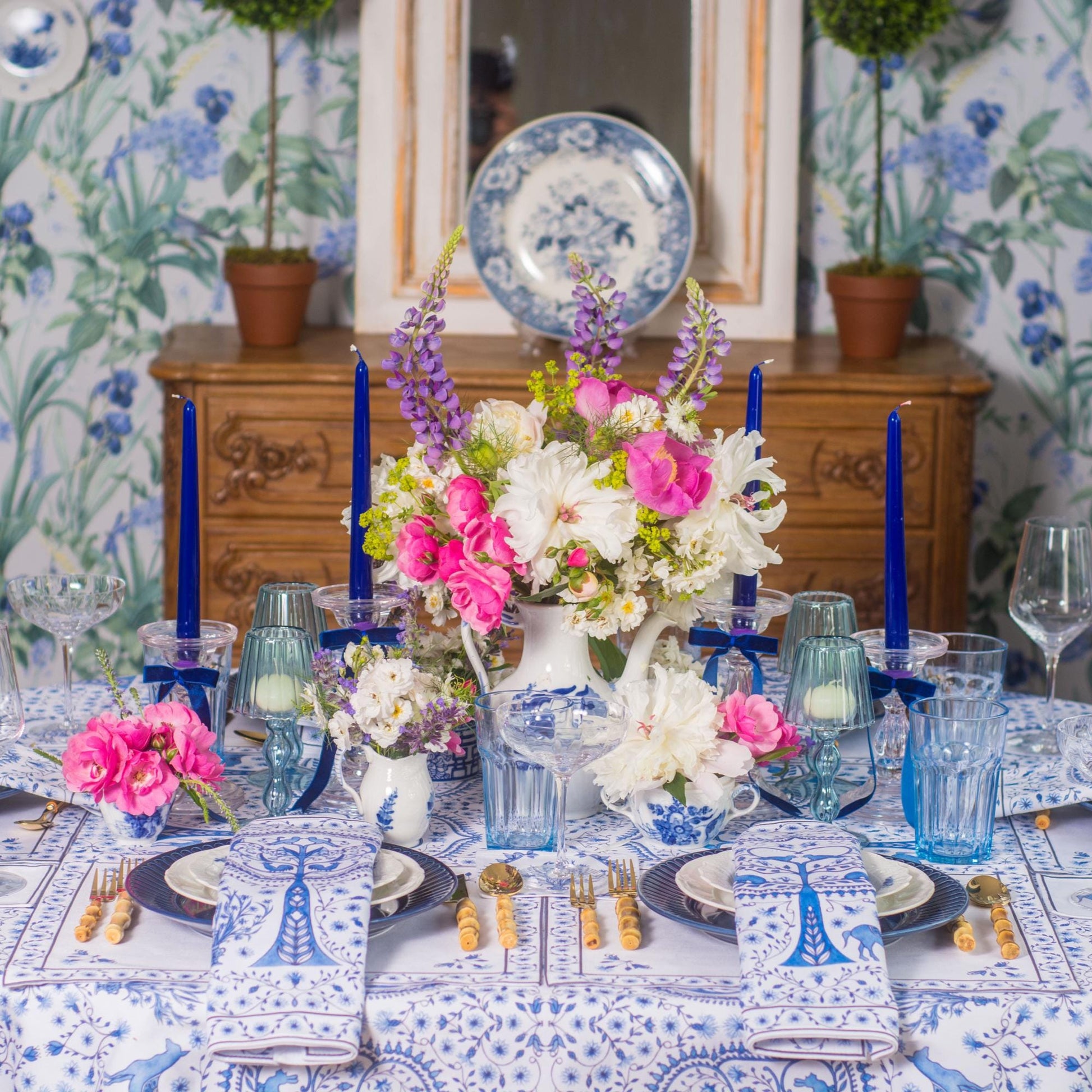Decorative table setting with cloth napkins, placemats and blue and white tablecloth, floral centerpieces and blue candles against a floral wallpapered wall.